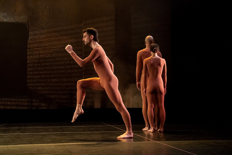 one dancer bearded in a rust colored unitard appears in profile prepared for a graceful run, in the back ground two other dancers in rust unitards face away from us and facing  the brick wall of the unadorned back stage at the Joyce.